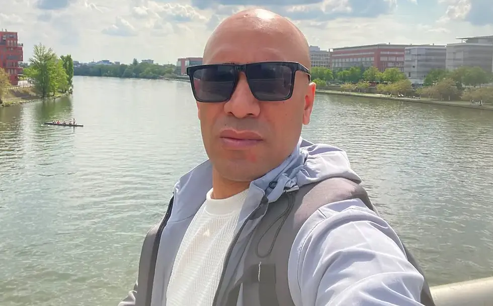 Chef Shree Ram Gyawali takes a selfie by a river, with city buildings, trees, and a small rowing boat in the sunny background.