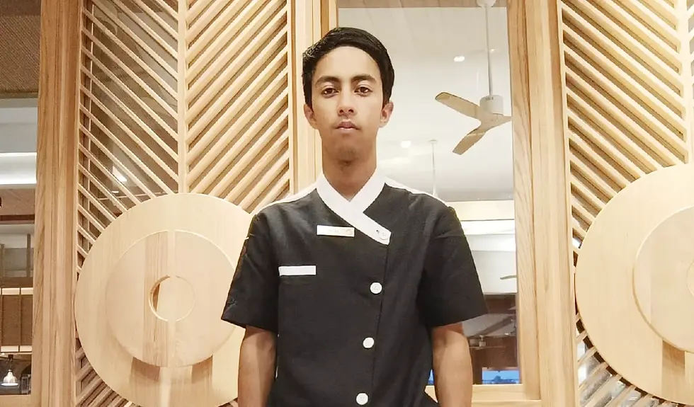 Chef Abish Chhetri stands indoors before wood slat decor and a ceiling fan, looking serious.