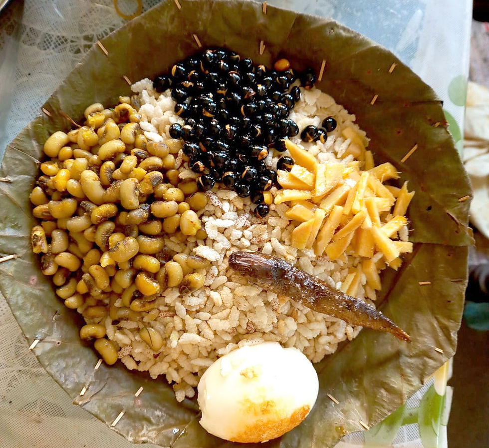 A leaf plate holds beans, puffed rice, a boiled egg, fried fish, and bamboo shoots. The setting is rustic with a lace tablecloth.