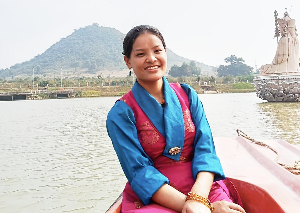 Chef Pasang Lhamu Sherpa in pink and blue dress sits on a boat. Background shows a large statue in water, with a distant hill and greenery.