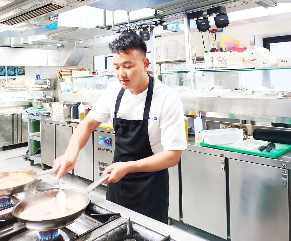 Chef Netra Magar in a black apron cooks in a bright, stainless steel kitchen, stirring sauce in a pan on a gas stove, focused and composed.