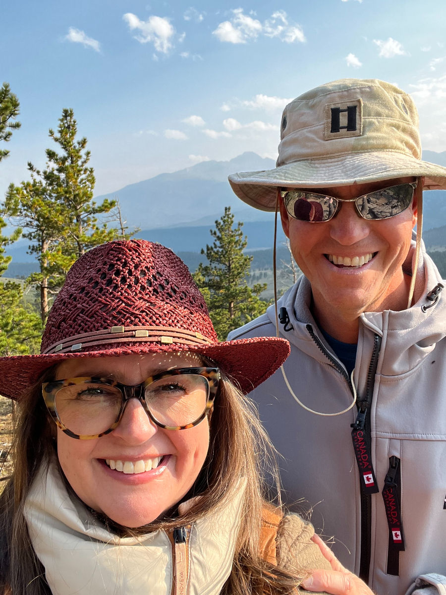 A smiling man and woman wearing hats, sunglasses, and jackets pose in a sunny, mountainous forest setting with a clear blue sky.