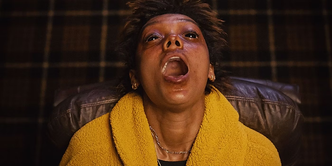 uma mulher, negra, cabelos curtos. está sentada em uma cadeira, com a cabeça completamente para trás, boca aberta e olhos quase completamente pretos. ela usa um roupão amarelo.