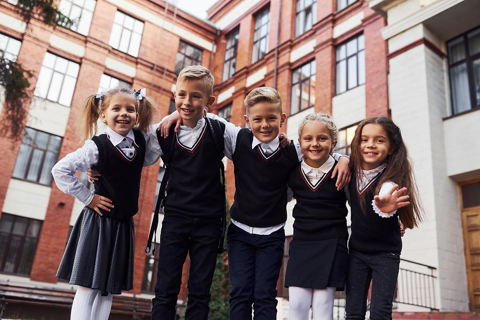 Five smiling schoolchildren in uniforms stand arm-in-arm outside a brick school building.