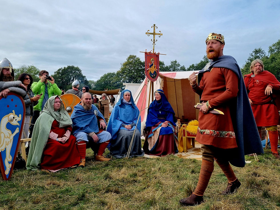 Medieval reenactors in colorful cloaks and armor gather by tents outdoors; a crowned King Harold Godwinson speaks while others listen and watch.