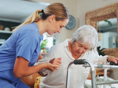 **Alt text:** *A physical therapist provides hands-on assistance to an older adult using a walker at home, focusing on safe mobility, balance, and functional independence.*