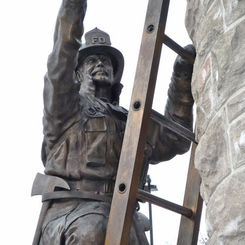 Bronze firefighter statue climbing a ladder against a stone wall. The statue wears a helmet with "FD" on it, conveying bravery.