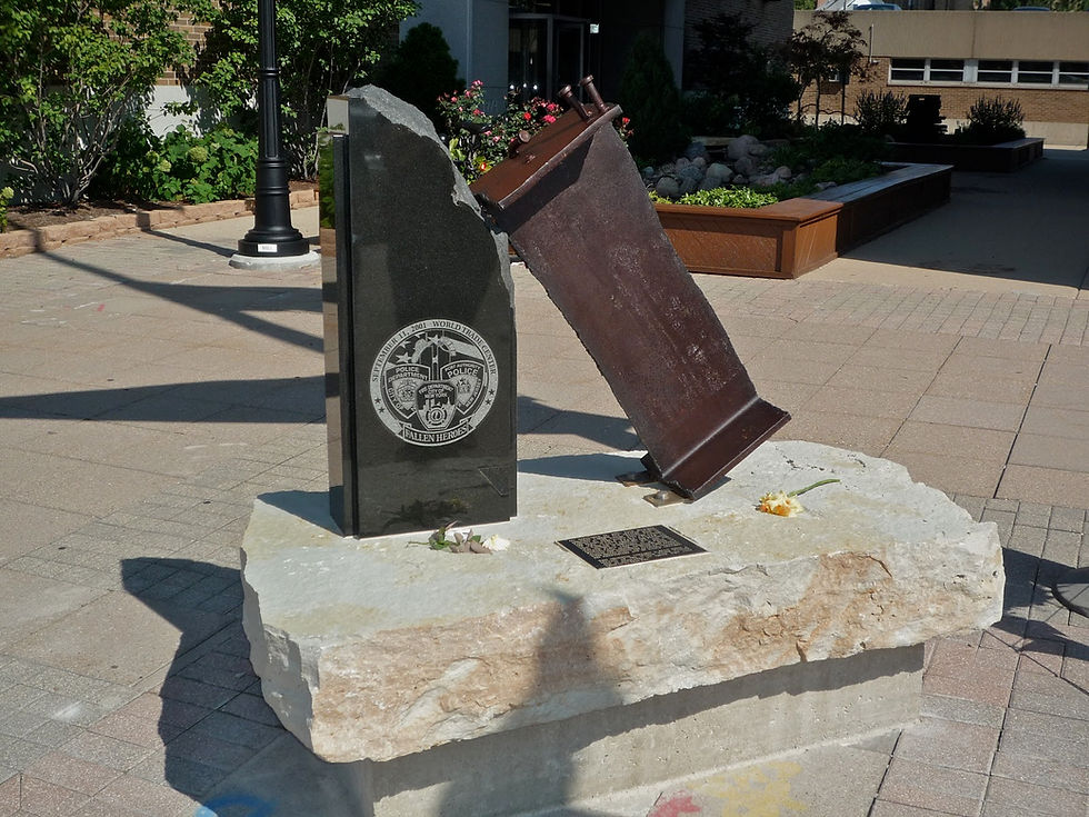 9/11 memorial sculpture featuring a steel beam and engraved stone. Plaque reads "Fallen Heroes." Flowers placed on stone, sunny day.