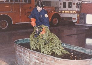 Firefighter in blue uniform demonstrates soaking a Christmas tree in a large metal tub. Fire trucks are visible in the background.