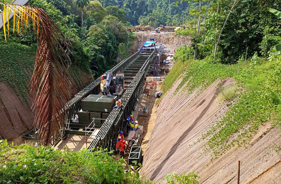 Bailey bridge construction in forested terrain, workers assembling modular steel framework with heavy equipment on steep excavation site.