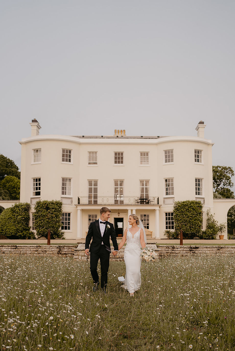 Bride and groom walking in long grass in front of rockbeare manor