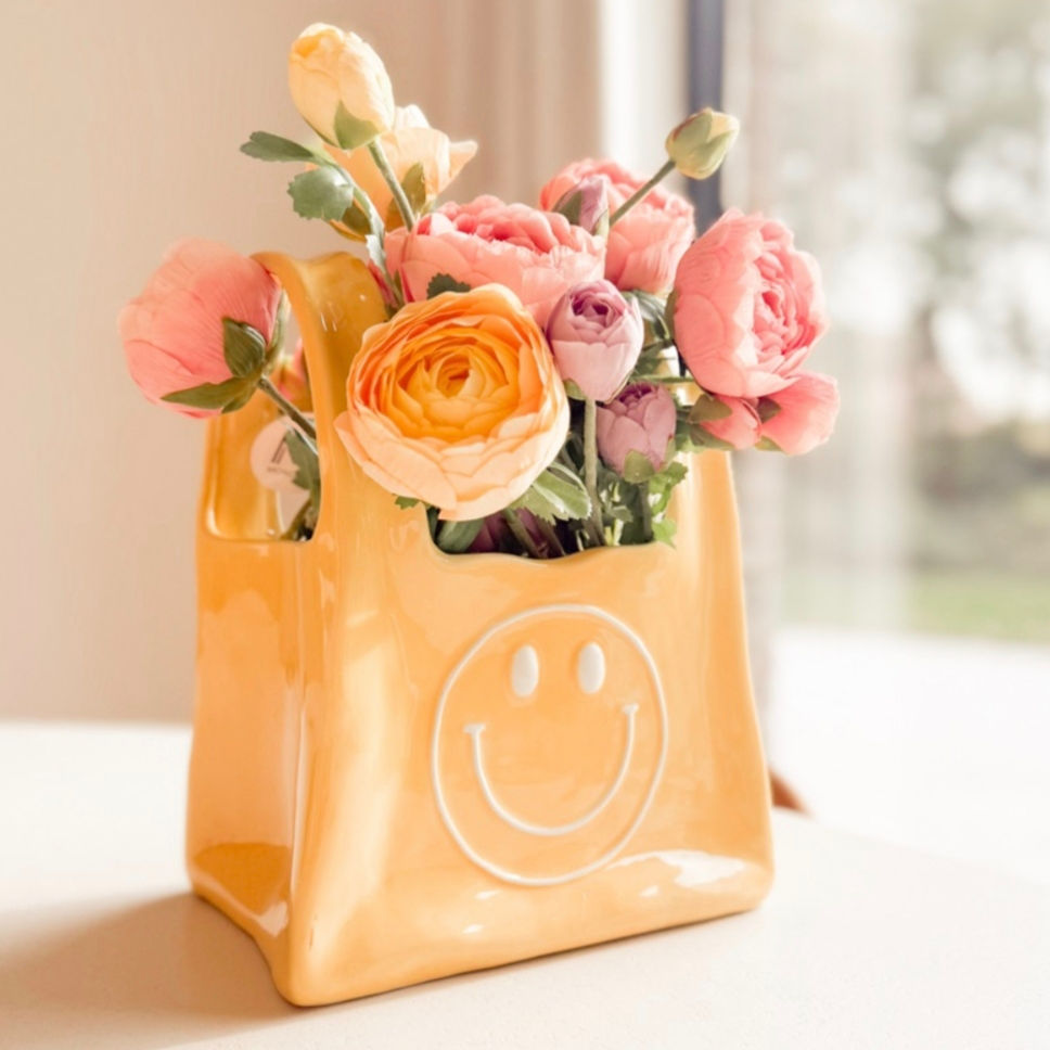 Miniature : Vue de profil, vase sac smiley emoji content en céramique jaune dans un décor intérieur avec fleurs.