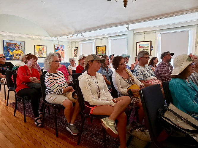 Rows of people sitting in a well lit room with pictures on the walls and a hardwood fllor listening attentively to a presentation