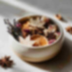 close-up of a ceramic bowl of natural potpourri with small air-dry clay leaf shapes, dried orange slices, rose petals, lavender buds, cinnamon sticks, and star anise, on a light marble surface