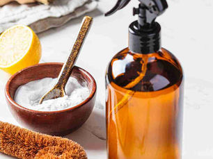 Natural cleaning essentials on a countertop: a brown glass spray bottle, a wooden bowl filled with baking soda and a golden spoon, a halved lemon, and eco-friendly cleaning brushes. Simmer & Stir branding visible.