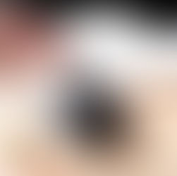 A hand is holding a dropper above a small gray bowl filled with smooth black stones, releasing a drop of liquid onto the stones. The bowl is placed on a round, dark stone base set on a wooden surface. In the foreground, a small amber glass bottle is visible, suggesting the use of essential oils or fragrance oils. A soft white fabric is blurred in the background, creating a calm, spa-like atmosphere.