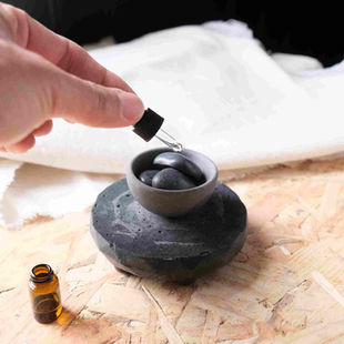 A hand is holding a dropper above a small gray bowl filled with smooth black stones, releasing a drop of liquid onto the stones. The bowl is placed on a round, dark stone base set on a wooden surface. In the foreground, a small amber glass bottle is visible, suggesting the use of essential oils or fragrance oils. A soft white fabric is blurred in the background, creating a calm, spa-like atmosphere.