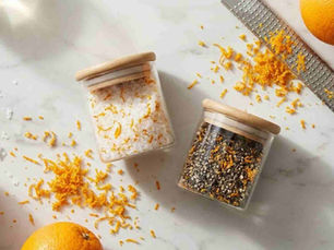 Two small clear glass jars with black or wooden lids, one filled with salt mixed with dried orange zest and the other with cracked black pepper mixed with dried orange zest. 