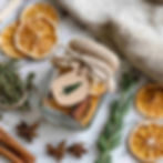 Flat lay of a natural, festive Dried Citrus & Spice Simmer Pot Kit on a white marble surface. A small clear glass jar filled with dried orange slices, cinnamon sticks, cloves, and rosemary sprigs, tied with jute twine and a kraft tag stamped with christmas tree.