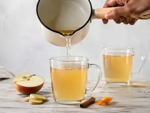Hand pouring a warm apple drink from a cream saucepan into a glass mug with apple, ginger, cinnamon and orange peel nearby.