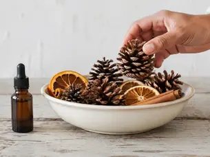 Hand legt een dennenappel in een keramieken schaal met gedroogde sinaasappel en kaneel naast een amberkleurig druppelflesje.