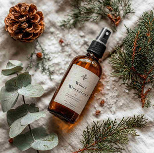 Flat lay of an amber glass spray bottle labelled “Winter Wonderland”, surrounded by pine needles, eucalyptus leaves, and peppermint sprigs. A few scattered snowflake-like salt crystals on a white linen background.