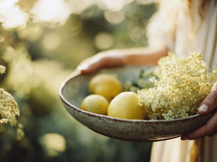 Woman holds a bowl of lemons and blossoms in a sunny garden. Soft light, earthy tones, and blurred greenery create a serene atmosphere.