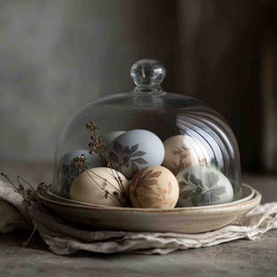 A moody, cinematic still life of naturally dyed Easter eggs with delicate botanical leaf prints, arranged loosely inside a shallow ceramic bowl and covered by a clear glass cloche. Eggs dyed in soft natural tones such as warm off-white, muted grey-blue, pale lavender and gentle beige, with organic leaf silhouettes visible on the shells.