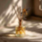 A minimalist scene with a homemade reed diffuser in a small clear or amber glass bottle, 5-6 reeds inserted. The bottle is filled with pale golden liquid. A few dried herbs (lavender, rosemary) float inside.