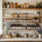 A beautifully styled home apothecary setup in a light, airy room with soft natural sunlight. Wooden shelves filled with labelled glass jars of dried herbs, mortar and pestle, amber bottles, fresh bundles of lavender and rosemary, clean minimal layout.