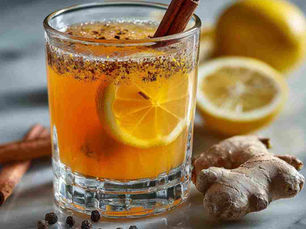 A minimalist close-up of a finished spice-infused mocktail served in a clear glass on a white marble kitchen worktop. The drink is golden-orange in colour with visible slice of lemon, cinnamon stick and fresh ginger floating gently.