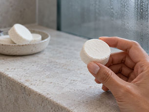 Hand holding a plain white DIY shower steamer above a ceramic dish on a stone bathroom ledge.
