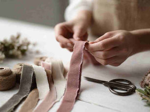 a white-grey Scandi tabletop. Velvet ribbons in muted sage, ivory, and dusty rose cut into equal strips lying on the side. Hands are tying a dusty rose ribbon strip onto a long, simple jute cord. A small pile of pre-cut velvet pieces on the side, along with scissors.