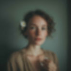 A woman with soft natural hair loosely tucked behind her ear, a small white jasmine flower placed delicately in her hair. She holds a minimal glass perfume bottle at chest height.