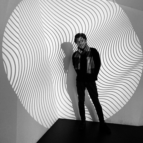 The image is of a man, the artist, dressed in black, shown full length in a casual pose near centre of frame shown at the corner of a room. The floor is in darkness and there is projected op-art light across the man and forming a circle of wavy black and white lines against the walls at the corner of the room.