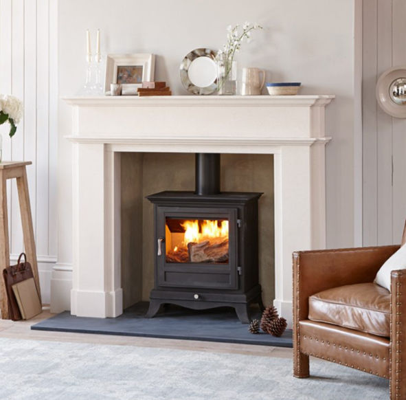 A cozy living room with a lit Chesneys Beaumont 8 black wood stove in a cream limestone fireplace. Brown leather chair and decor on the mantel create a warm ambiance.