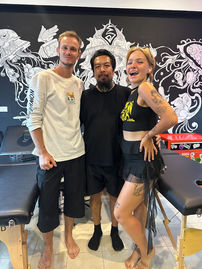 Three people pose for photo in a tattoo studio, smiling. Tattoo artwork background.