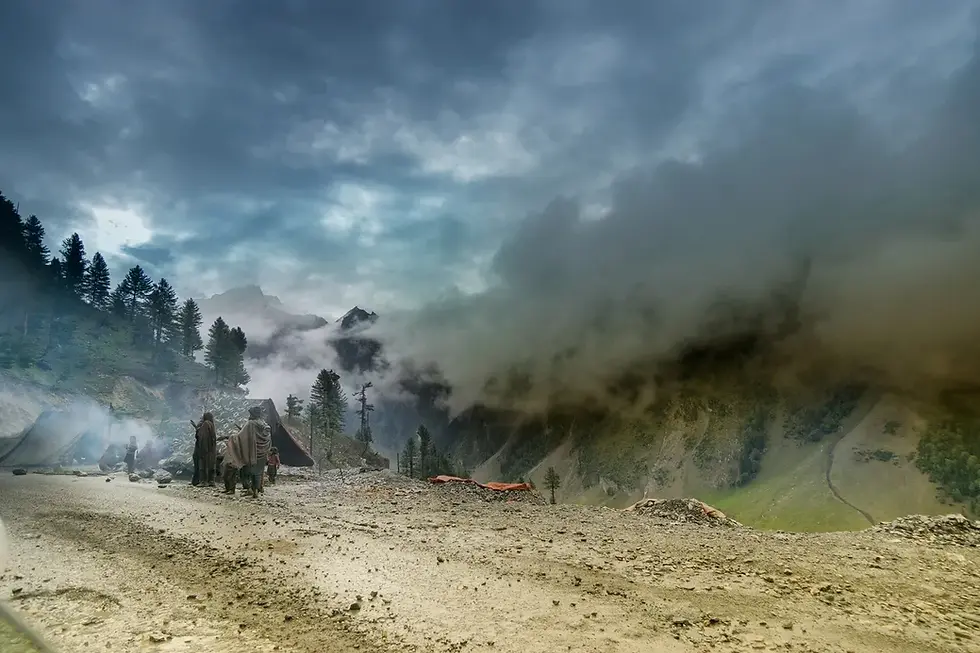 People in cloaks walk along a rugged path amidst misty mountains and pine trees, under a cloudy sky, creating a serene atmosphere.