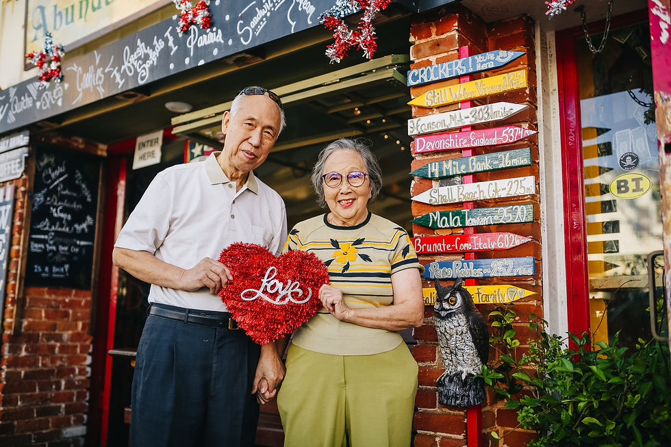 A happy, proud older Asian couple holds a red heart in from of their colorful shop.