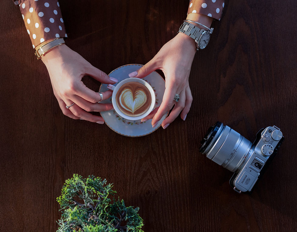 A woman's hands hold a latte with a heart on top. She has a camera next to her on the table.