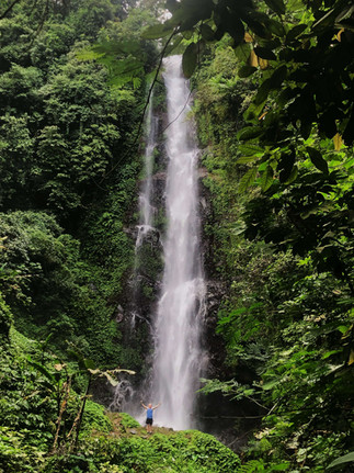 Mike standing at the foot of melanting falls with his hands up in the air, looking so small compared to the huge wall of water