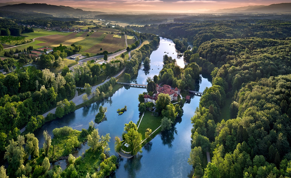 Aerial view of a river winding through forests and fields, with a red-roofed castle on an island at sunset in Slovenia
