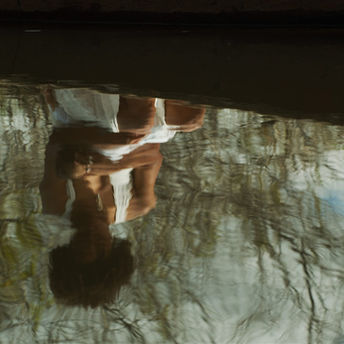 A cineasta Safira Moreira refletida nas águas do rio Paraguaçu em cena do seu filme Cais, produção baiana que foi a grande vencedora do 14º Olhar de Cinema. Na imagem, está o reflexo um pouco difuso e de ponta cabeça na água de uma mulher negra sentada com um vestido branco e ramos de árvores ao fundo dela.
