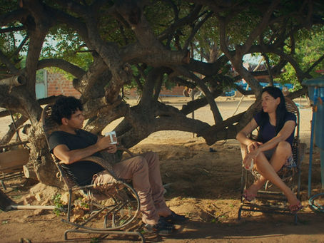 João Vieira Torres e a prima em cena do seu filme Aurora (2025). Na imagem, um homem e uma mulher estão sentados em cadeiras de balanço de palha à frente de uma árvore de troncos e galhos laterais e copa baixa, mas bem horizontalizada. Sentado de lado para a câmera, o homem negro com cabelos cacheados veste uma camiseta preta, calça marrom e tênis preto, além de segurar uma caneca branca na mão direita. De frente para a câmera, está sentada a mulher parda, cabelos pretos longos e lisos, bermuda, chinelo de tira rosa e também segura uma caneca branca na mão esquerda. Ao lado esquerdo dela, há uma grande caixa d'água azul.