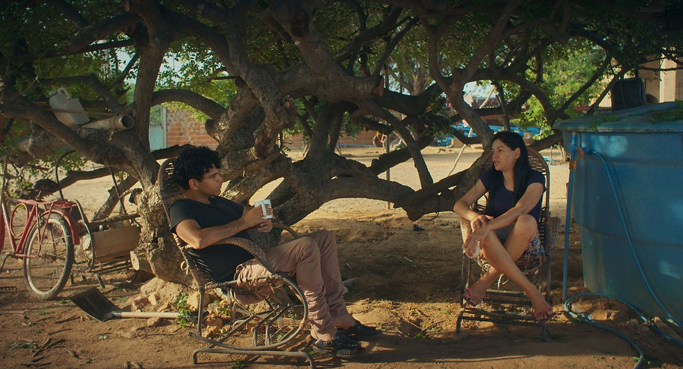 João Vieira Torres e a prima em cena do seu filme Aurora (2025). Na imagem, um homem e uma mulher estão sentados em cadeiras de balanço de palha à frente de uma árvore de troncos e galhos laterais e copa baixa, mas bem horizontalizada. Sentado de lado para a câmera, o homem negro com cabelos cacheados veste uma camiseta preta, calça marrom e tênis preto, além de segurar uma caneca branca na mão direita. De frente para a câmera, está sentada a mulher parda, cabelos pretos longos e lisos, bermuda, chinelo de tira rosa e também segura uma caneca branca na mão esquerda. Ao lado esquerdo dela, há uma grande caixa d'água azul.