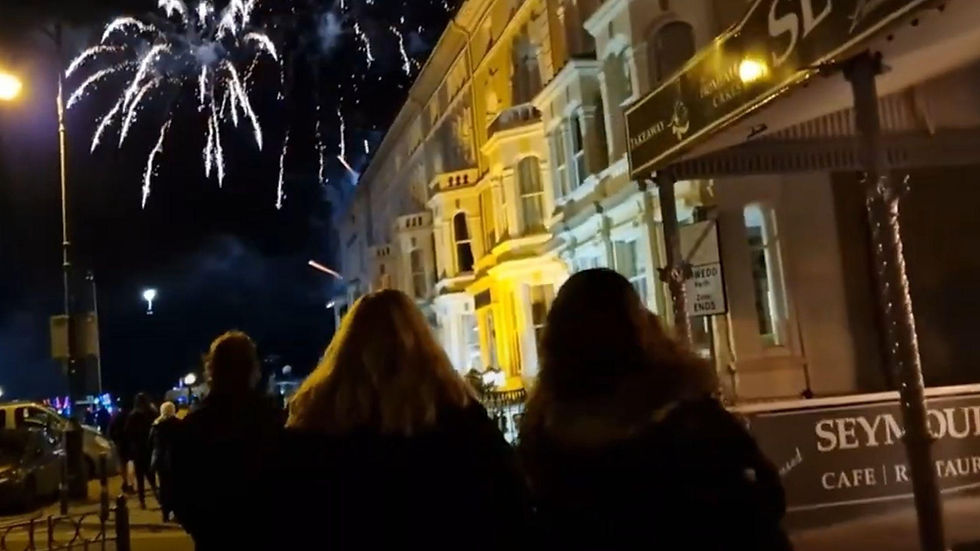 Three people watch fireworks over lit buildings on a night street, with a Seymour cafe sign in view by Llandudno Great Escapes