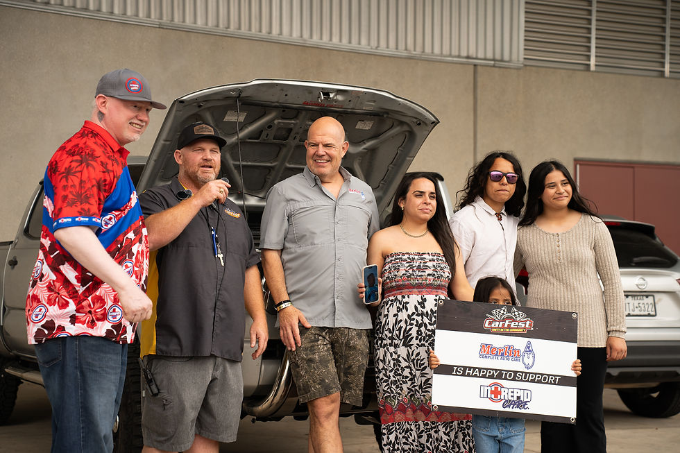 Group poses beside an open car hood at CarFest, smiling and holding a Merlin/Intrepid Care support sign.