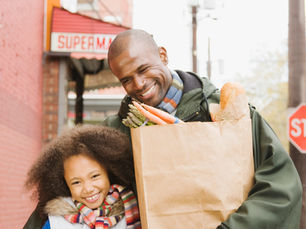 Young black father with his young daughter walking down a street laughing and smiling as if having a fun conversation. They are wearing wintery clothing and he is carrying a brown paper sack full of groceries as if they are on their way home from the supermarket seen in the background