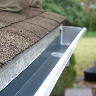 A close-up of a clean, freshly installed house gutter along a roof with brown shingles. Th