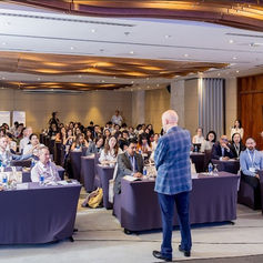 A speaker in a blue plaid jacket addresses a packed conference room of seated delegates from various international organizations, with roll-up banners and a camera operator visible in the background.